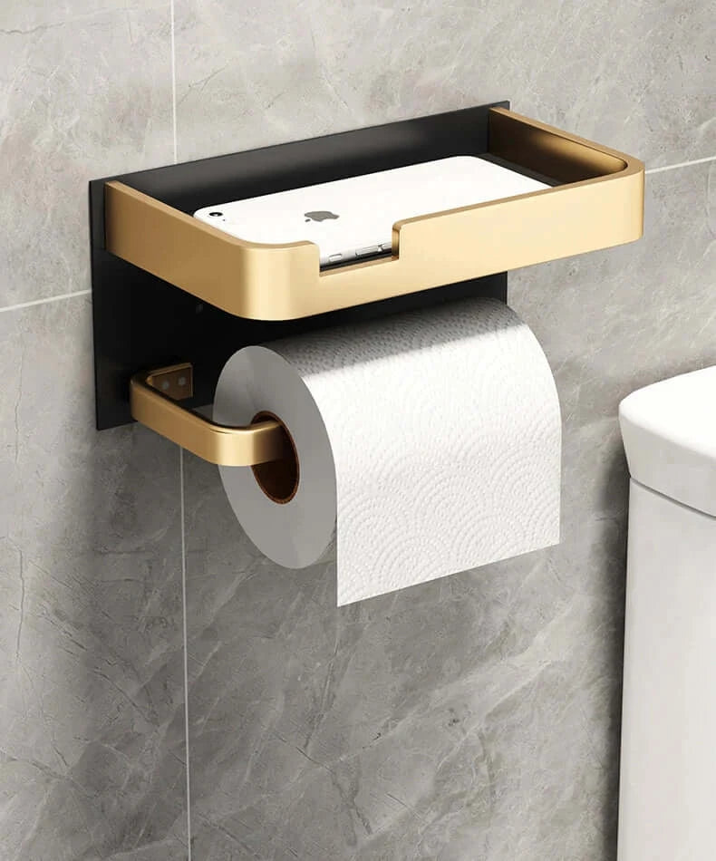 Wall-mounted Toilet Paper Roll Holder Phone Shelf in gold and black, holding a smartphone above the toilet paper roll.